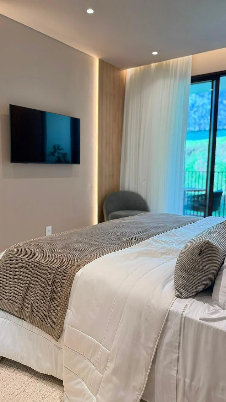 Suíte master com cama, poltrona e porta de vidro aberta para a varanda